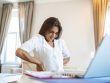 Portrait of young stressed woman sitting at home office desk in front of laptop, touching aching back with pained expression, suffering from backache after working on laptop