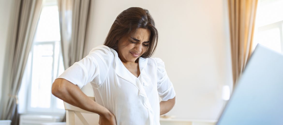 Portrait of young stressed woman sitting at home office desk in front of laptop, touching aching back with pained expression, suffering from backache after working on laptop