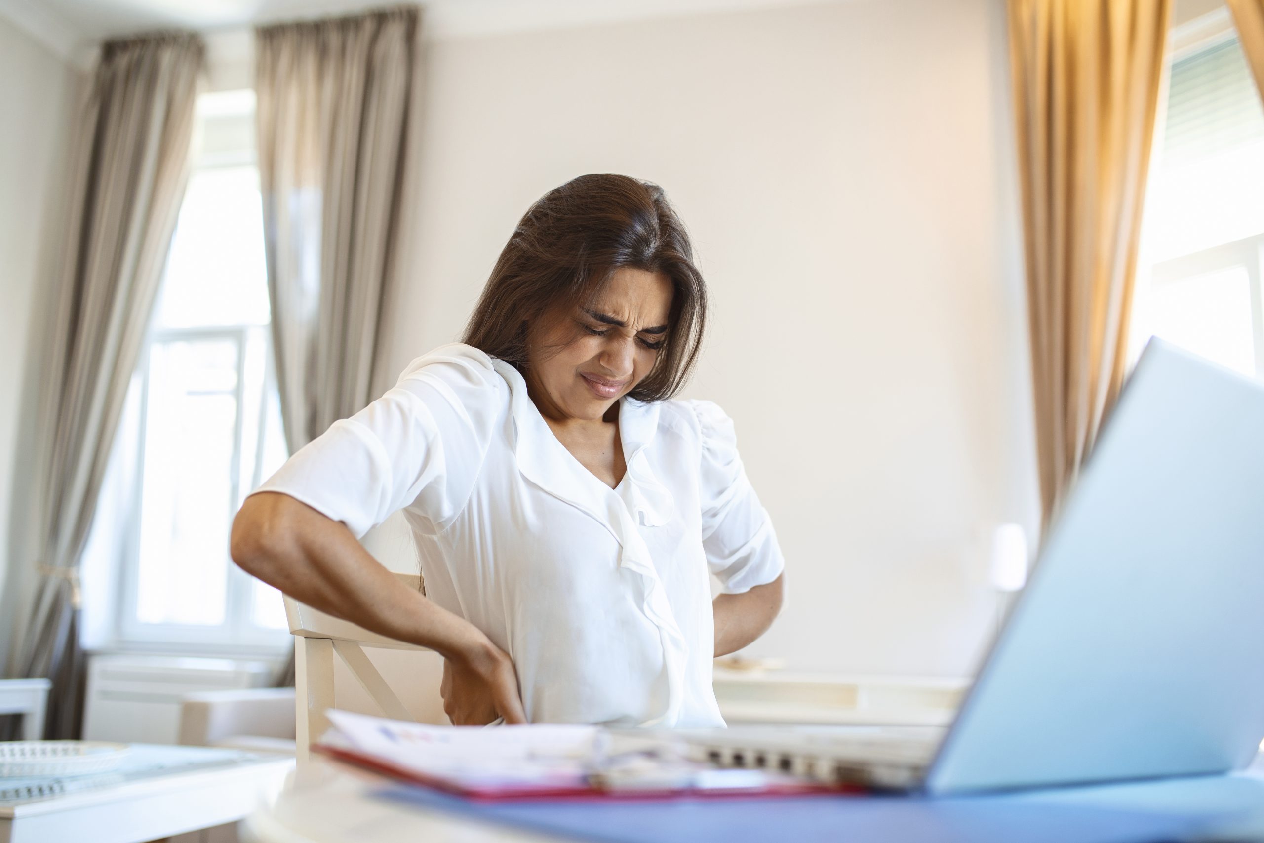 Portrait of young stressed woman sitting at home office desk in front of laptop, touching aching back with pained expression, suffering from backache after working on laptop